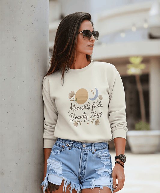 A woman in sunglasses leaning against a pillar in usa wearing a cream AWonders sweatshirt featuring a celestial design and the quote "Moments fade, Beauty stays."